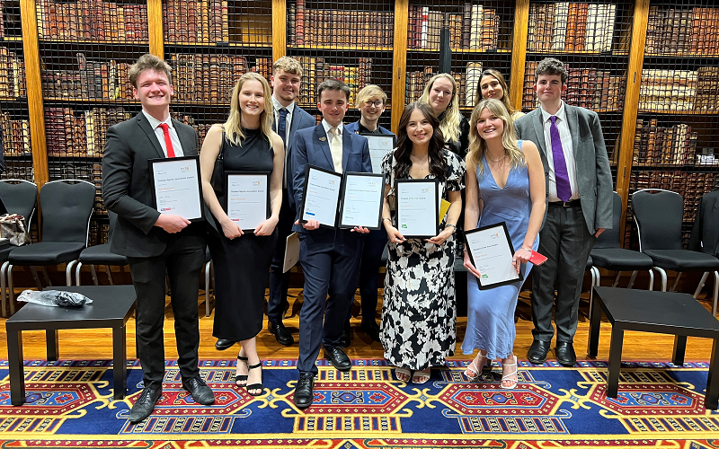 All of our News Associates grads shortlisted for the NCTJ Awards for Excellence standing and holding their certificates and smiling. They are all smartly dressed and standing in front of rows of books on a shelf in the library at the Royal College of Physicians.