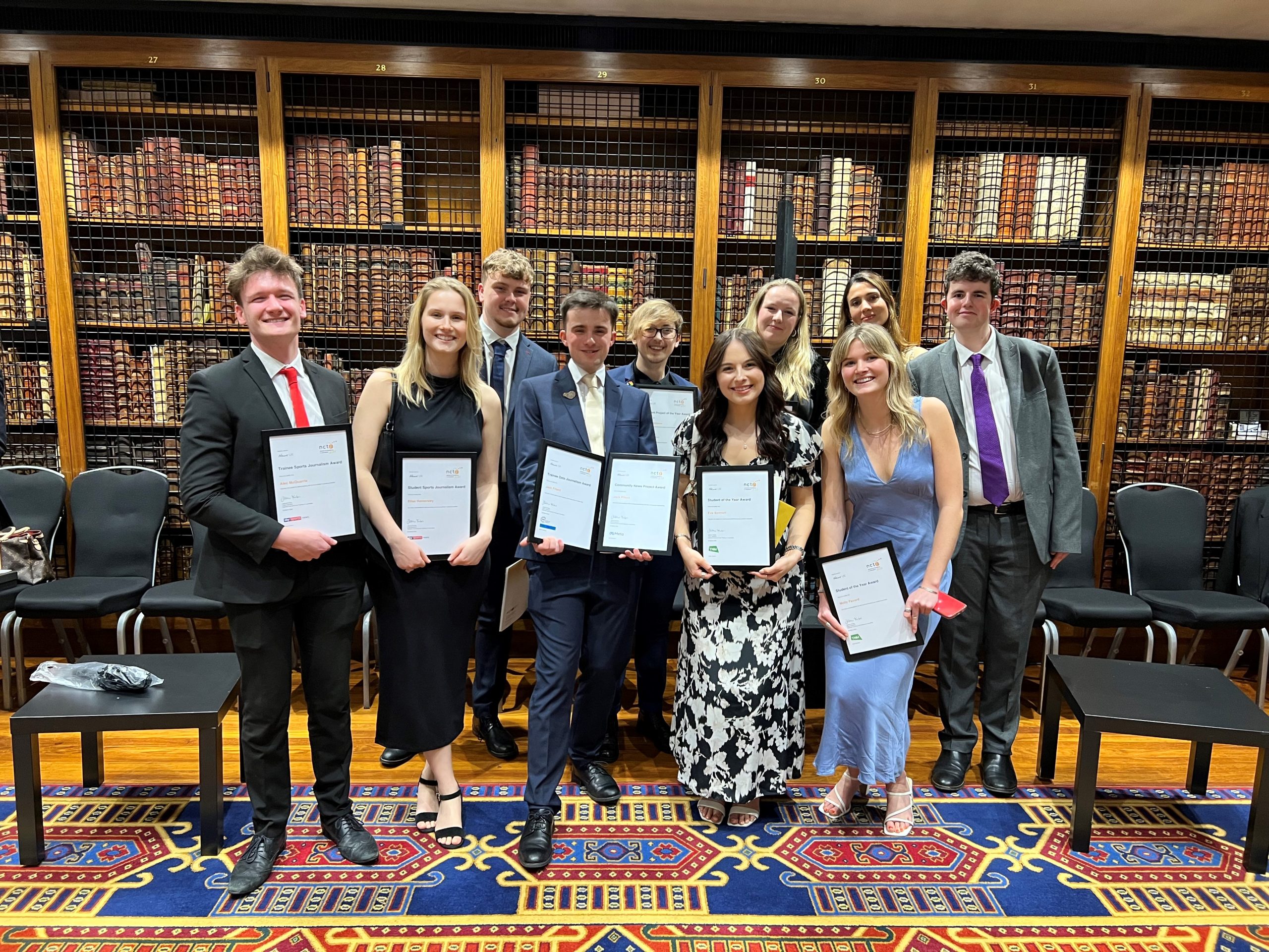 All of our News Associates grads shortlisted for the NCTJ Awards for Excellence standing and holding their certificates and smiling. They are all smartly dressed and standing in front of rows of books on a shelf in the library at the Royal College of Physicians.