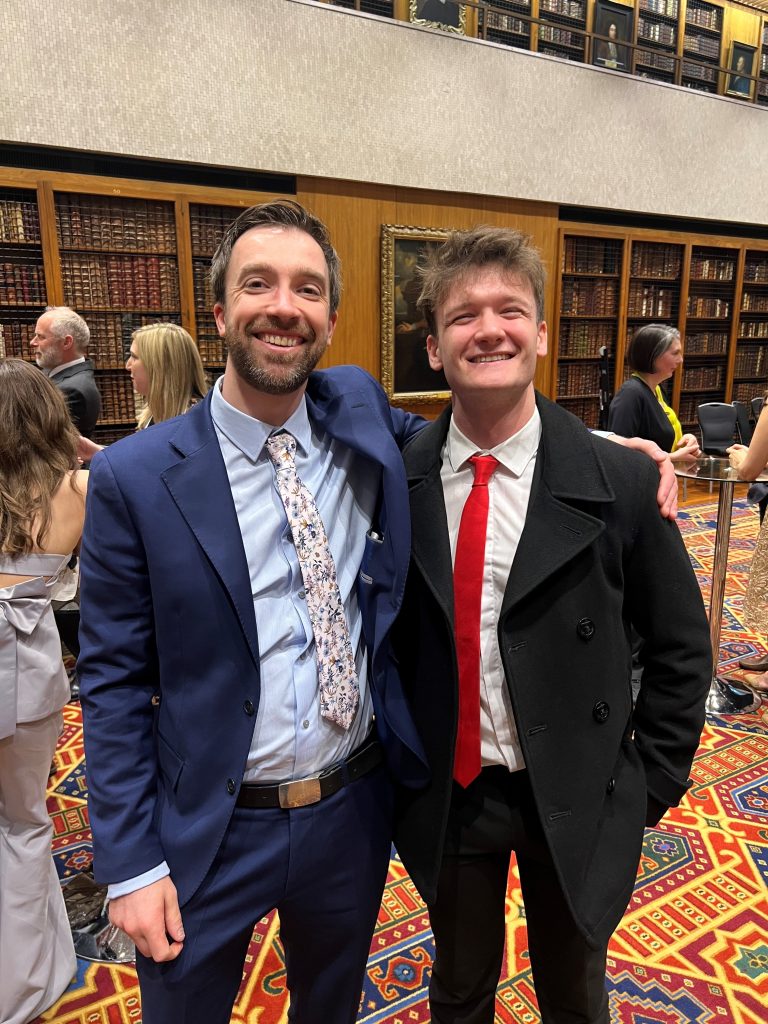 News Associates course director Graham Moody and graduate Alec McQuarrie. They are both smiling and smartly dressed and standing in front of rows of books on a shelf in the library at the Royal College of Physicians at the NCTJ Awards for Excellence 2023.