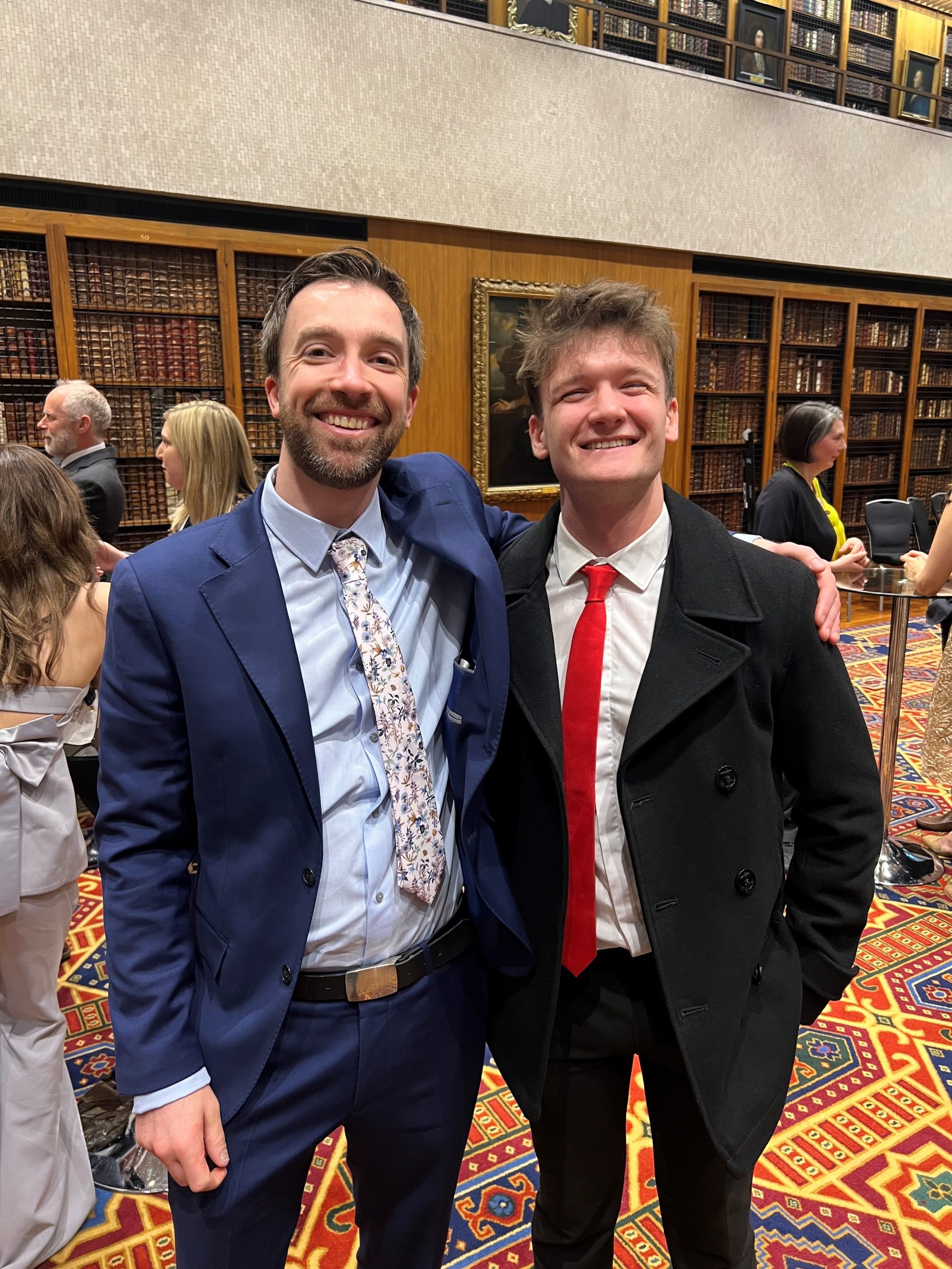 News Associates course director Graham Moody and graduate Alec McQuarrie at the NCTJ Awards for Excellence 2023. They are both smiling and smartly dressed and standing in front of rows of books on a shelf in the library at the Royal College of Physicians.