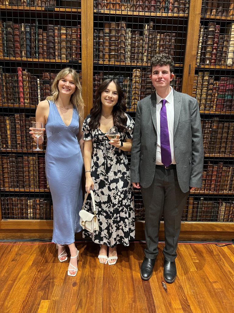 News Associates graduates Molly Pavord, Eve Bennett and Noah Keate at the NCTJ Awards for Excellence 2023. They are all smiling and smartly dressed and standing in front of rows of books on a shelf in the library at the Royal College of Physicians.