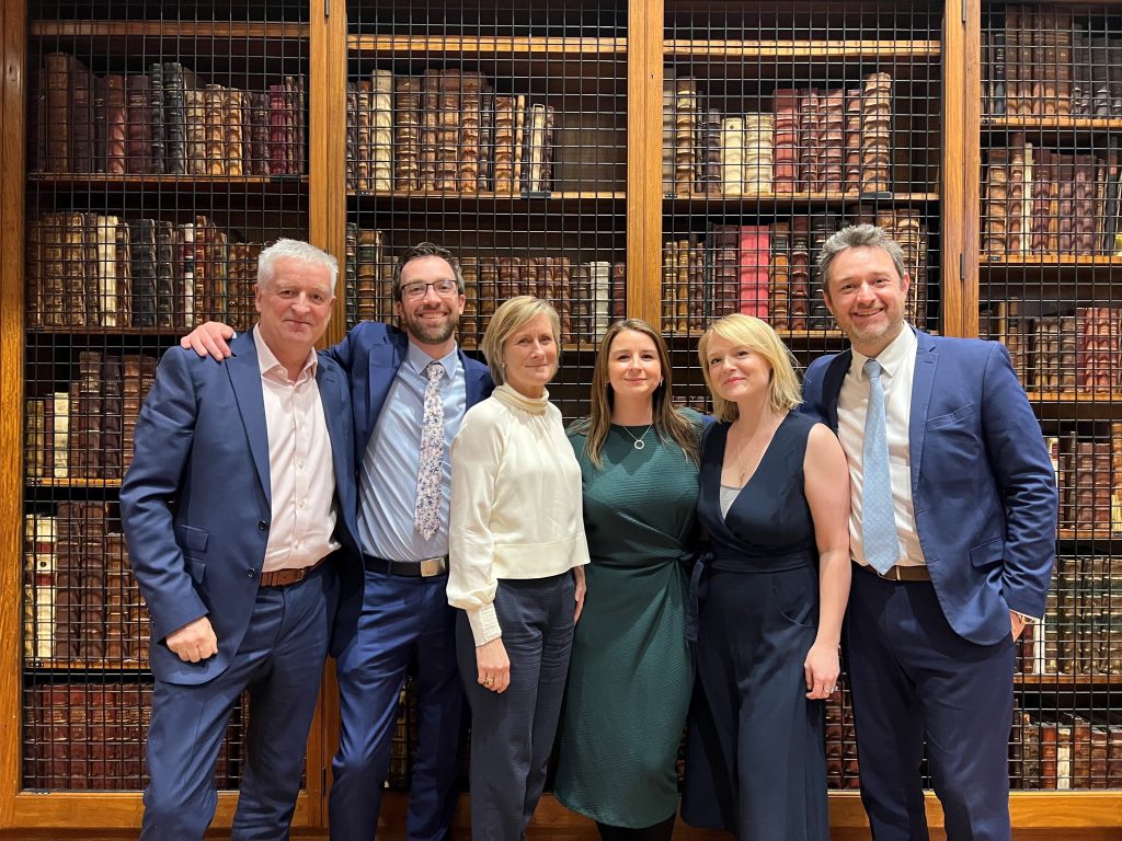 News Associates staff Graham Dudman, Graham Moody, Angela Catto, Lucy Dyer, Rachel Bull and James Toney at the NCTJ Awards for Excellence.  They are all smartly dressed and standing in front of rows of books on a shelf in the library at the Royal College of Physicians.