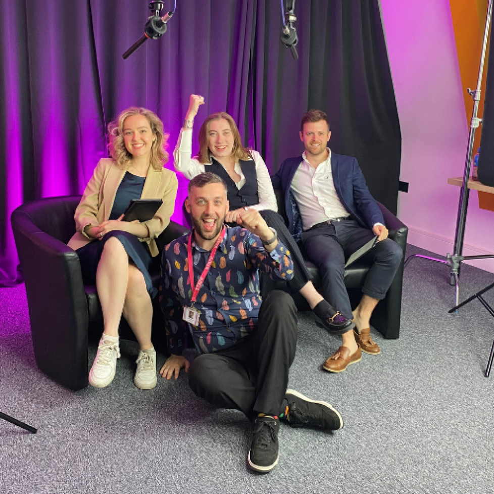 Our three trainees and journalism tutor who produced our general election livestream through the night. They are all sitting and cheering, celebrating the end of a successful night of coverage. They are all dressed smartly sitting in front of a black back drop with purple lighting.