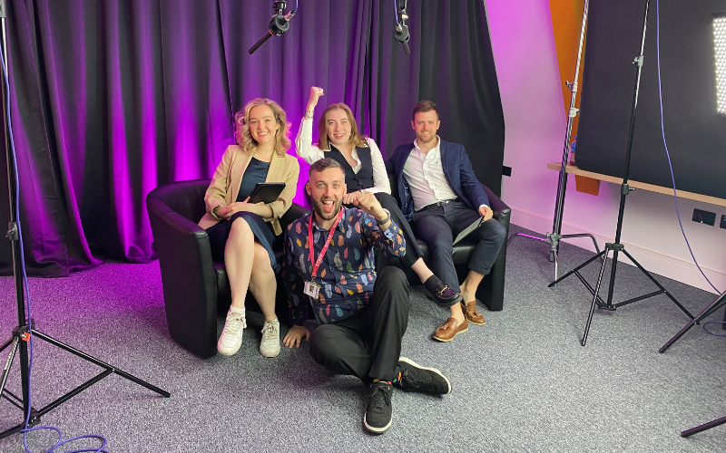 Our three trainees and journalism tutor who produced our general election livestream through the night. They are all sitting and cheering, celebrating the end of a successful night of coverage. They are all dressed smartly sitting in front of a black back drop with purple lighting.