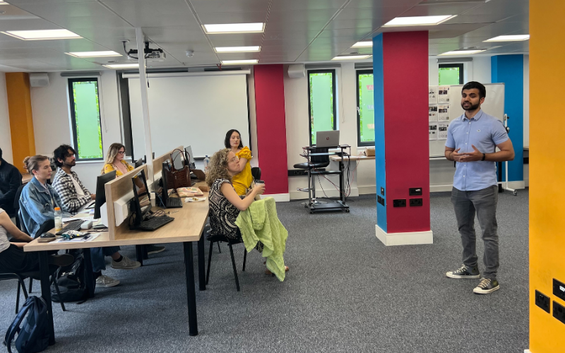 News Associates graduate Kumail Jaffer (pictured on the right-hand side in a blue polo) giving a talk to a group of News Associates trainees who attentively listen to his session at their desks in the News Associates newsroom that is fitted with multicoloured bollards.