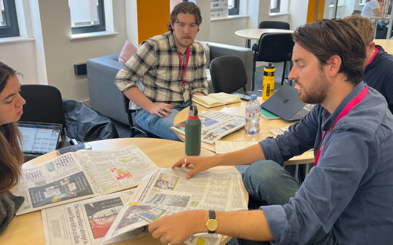 A group of four News Associates trainees all with a newspaper to hand discuss the news in preparation for their paper reviews. They are sat at two circular tables that have been moved together and also have their laptops, notebooks and water bottles on the tables.