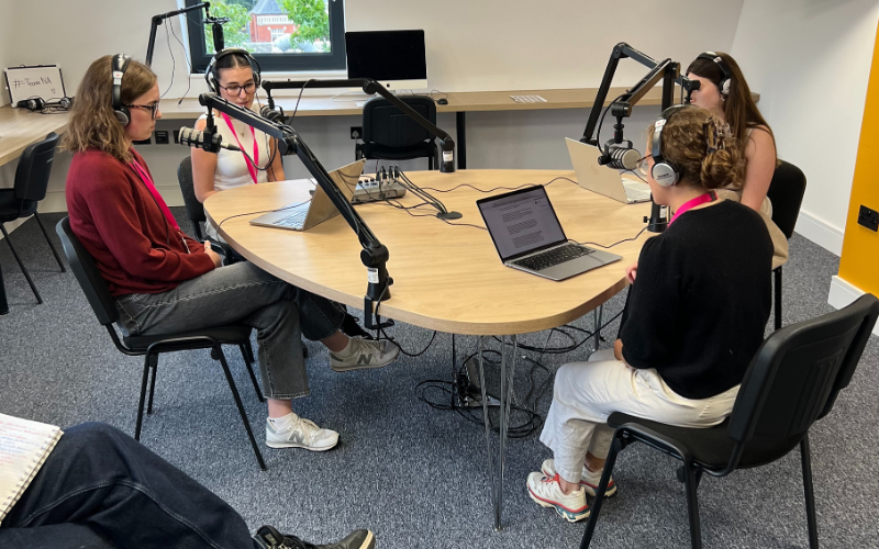 Four News Associates trainees are sat at desk recording their own radio news bulletins. Their all have their microphones and headphones set up and are reading their scripts from their laptops in a broadcast studio.