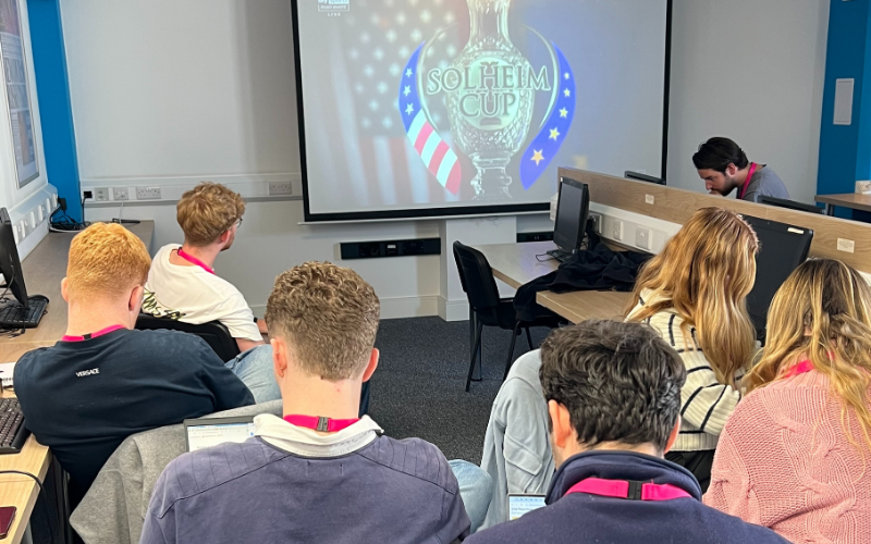 News Associates trainees watching the Solheim Cup on a projector in a classroom with laptops to hand.