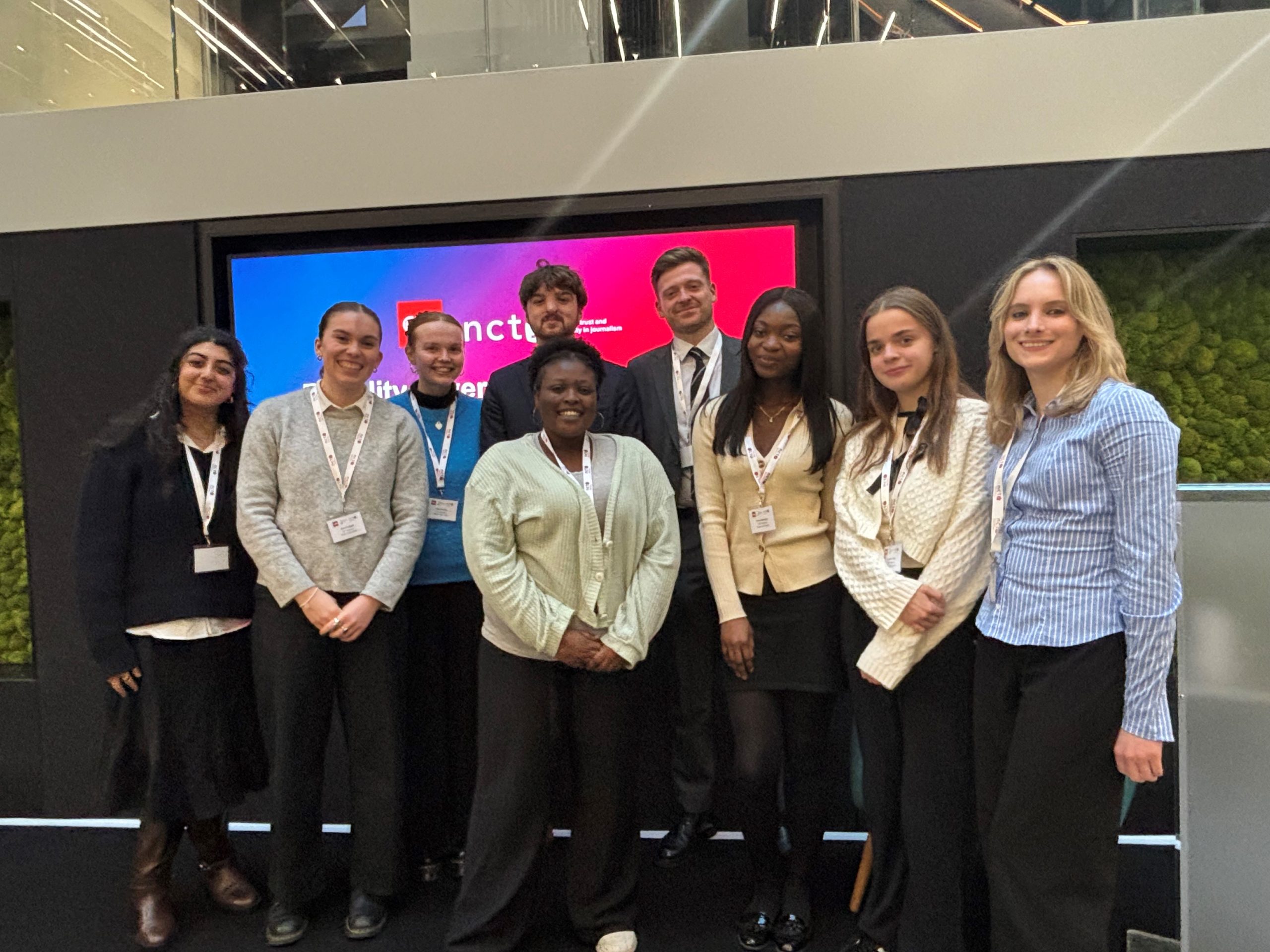 Nine people dressed in formalwear and lanyards stand together on a stage in front of a colourful screen which includes the NCTJ logo. They are all smiling together at the camera. 