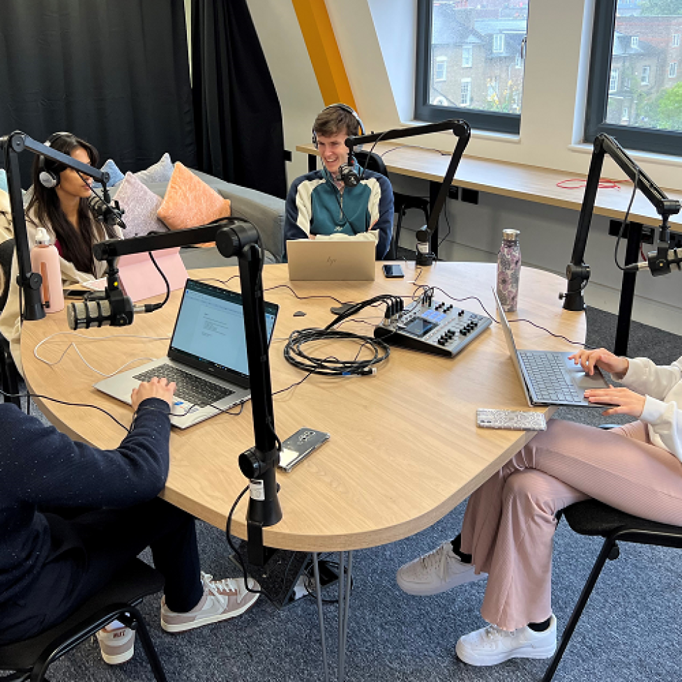 Four News Associates trainees sitting around a desk with mics and laptops recording a podcast in our studio.
