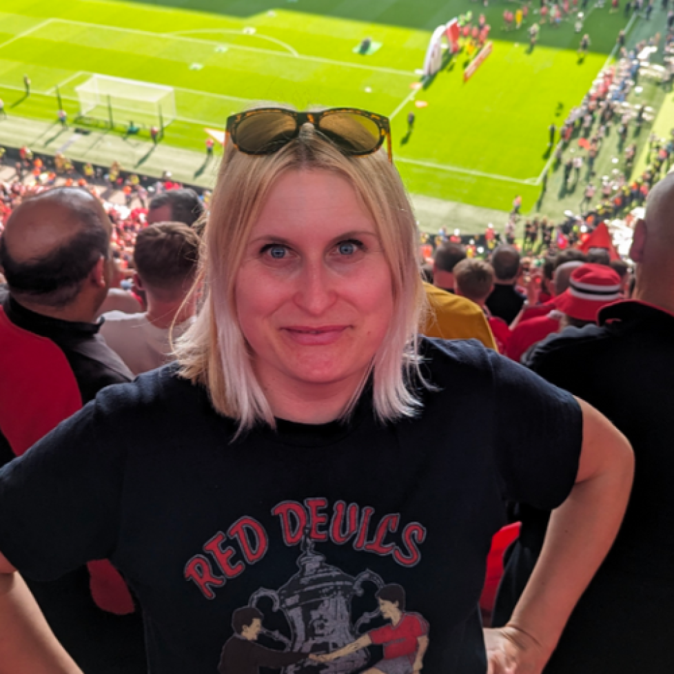 FWA Vikki Orvice Student Football Writer of the Year, Carly, with short blonde hair and sunglasses on her head wearing a black Red Devils t-shirt standing with her hands on her hips smiling up at the camera from the stands of a stadium with the pitch behind her.