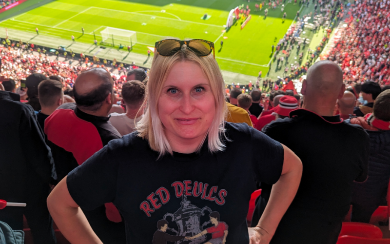 FWA Vikki Orvice Student Football Writer of the Year, Carly, with short blonde hair and sunglasses on her head wearing a black Red Devils t-shirt standing with her hands on her hips smiling up at the camera from the stands of a stadium with the pitch behind her.