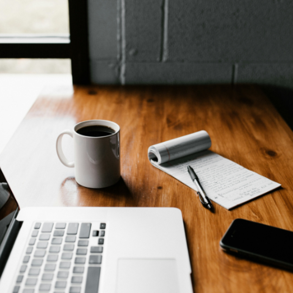 A photo of a desk with a laptop, notebook and cup of coffee on. The image is being used to illustrate an article titled How to prepare for a News Associates course.